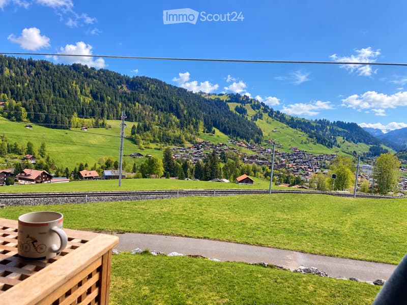 Balcony with a wooden table and a cup, overlooking a scenic valley with a village, green hills, and mountains.