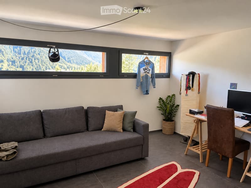 A cozy living room with a grey sofa, wooden desk, chair, and a potted plant. The room features large windows with a scenic mountain view and a hanging jersey on the wall.