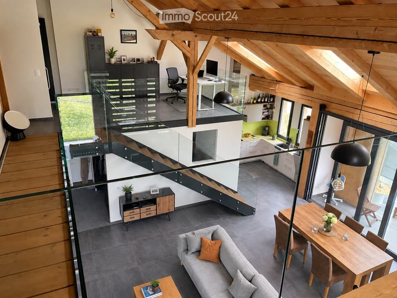 Open-concept living and dining area with exposed wooden beams, modern furniture including a sofa, coffee table, dining table, and office area with a desk and chair. The space features a glass staircase and large windows offering natural light.