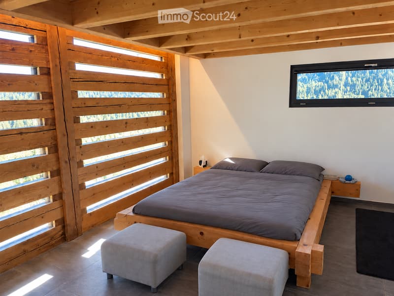 Modern bedroom with wooden bed, nightstand, two grey stools, and a wall-mounted screen displaying a scenic view.