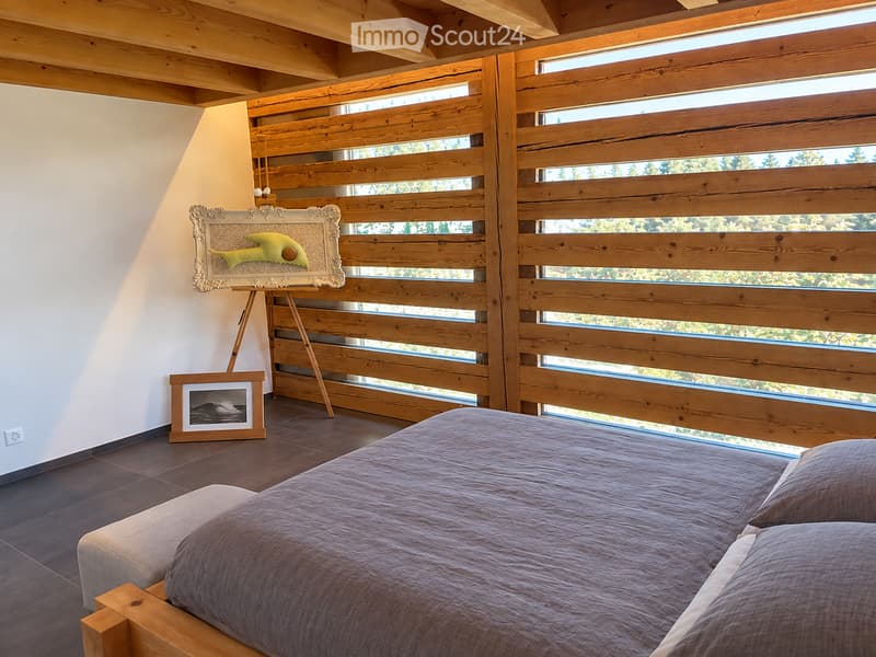 Modern bedroom with a wooden slatted wall, large windows, a bed with pillows, picture frames, and an easel with artwork.