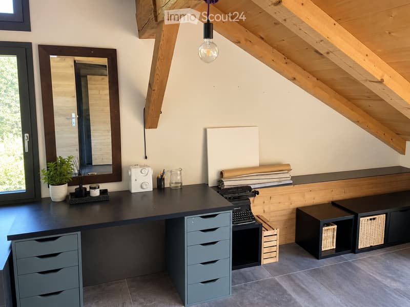 A modern, well-lit workspace with a large wooden desk, a mirror, a potted plant, a typewriter, storage drawers, and wicker baskets. The room features exposed wooden beams and a slanted ceiling.