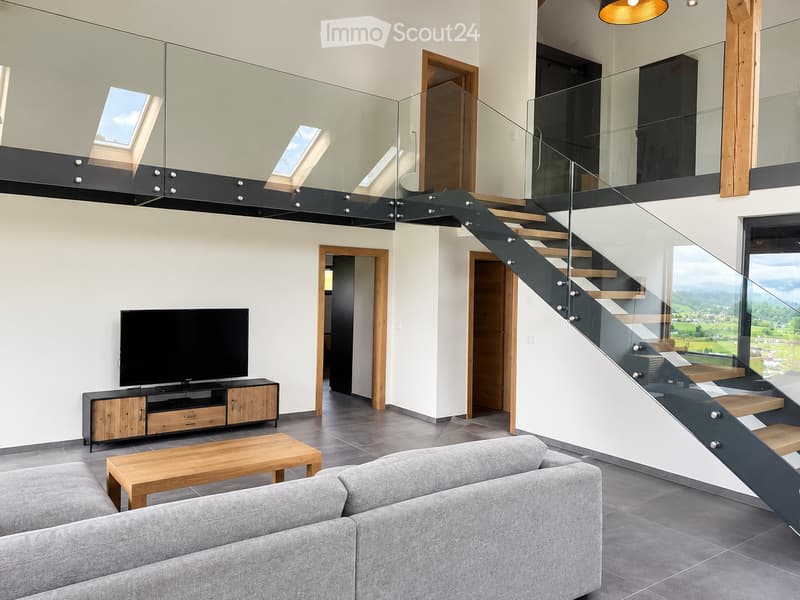 Modern living room with grey sofa, wooden coffee table, TV on a wooden stand, and a staircase leading to an upper level with glass railings and skylights.