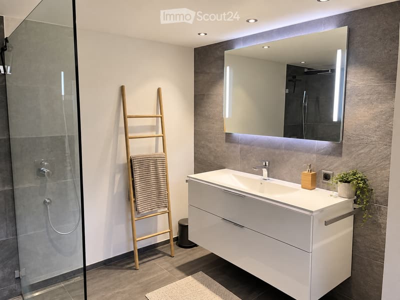 Modern bathroom with a white vanity, large mirror, glass shower enclosure, and a wooden towel ladder with a towel.