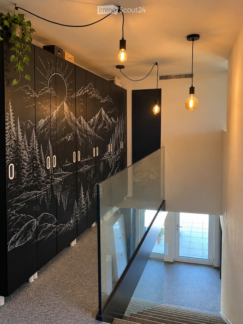 Modern hallway with a glass staircase, black wall mural depicting mountains and trees, pendant lights, and a small plant on the wall.