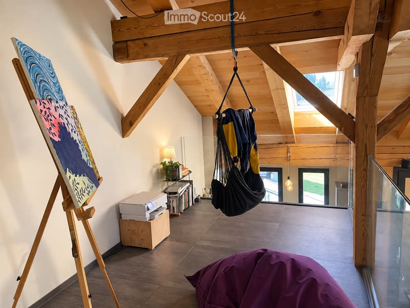 A spacious, modern room with exposed wooden beams and large windows. Features include an easel with artwork, a bean bag chair, a hanging clothes hanger, a small table with a lamp and potted plant, and a printer on a wooden stand.