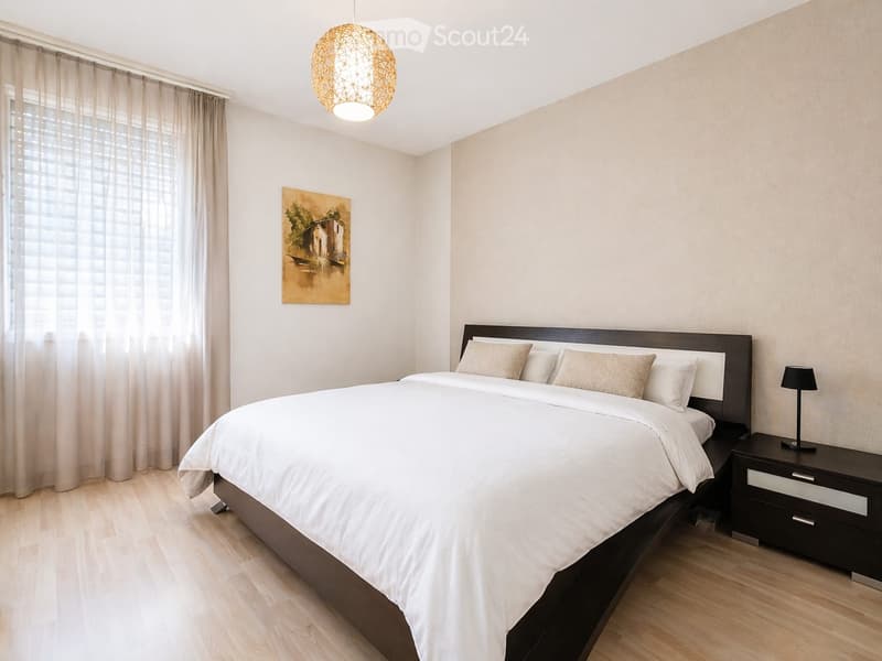 Modern bedroom with a large bed, nightstand, lamp, and a painting on the wall. The room features light-colored curtains, a pendant light fixture, and wooden flooring.