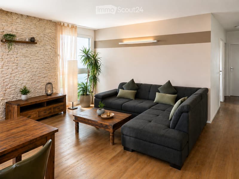 Modern living room with a dark grey sectional sofa, wooden coffee table, dining table, and various decorative items including plants and lamps.
