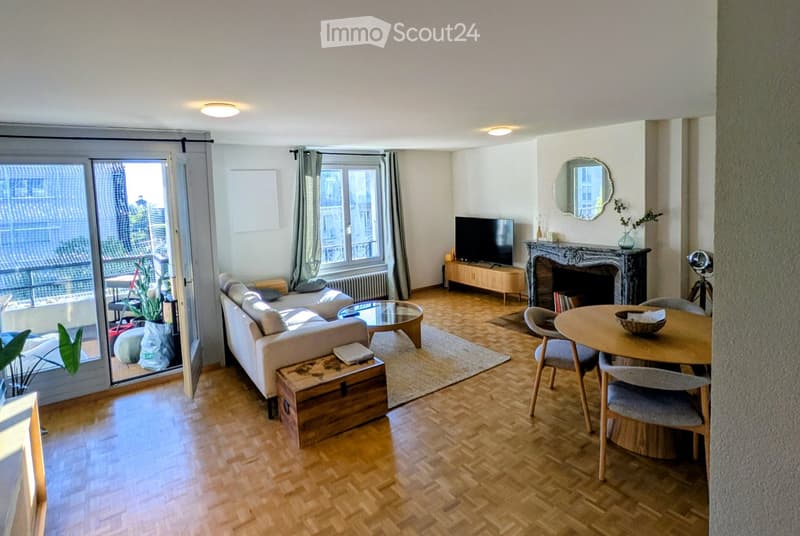 Bright living room with wooden flooring, large windows, balcony access, sofa, coffee table, dining table, fireplace, and decorative items.