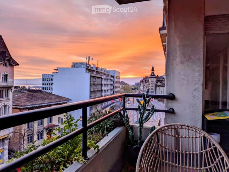 Balcony with a view of the city at sunset, featuring a wicker chair, potted plants, and a metal railing.