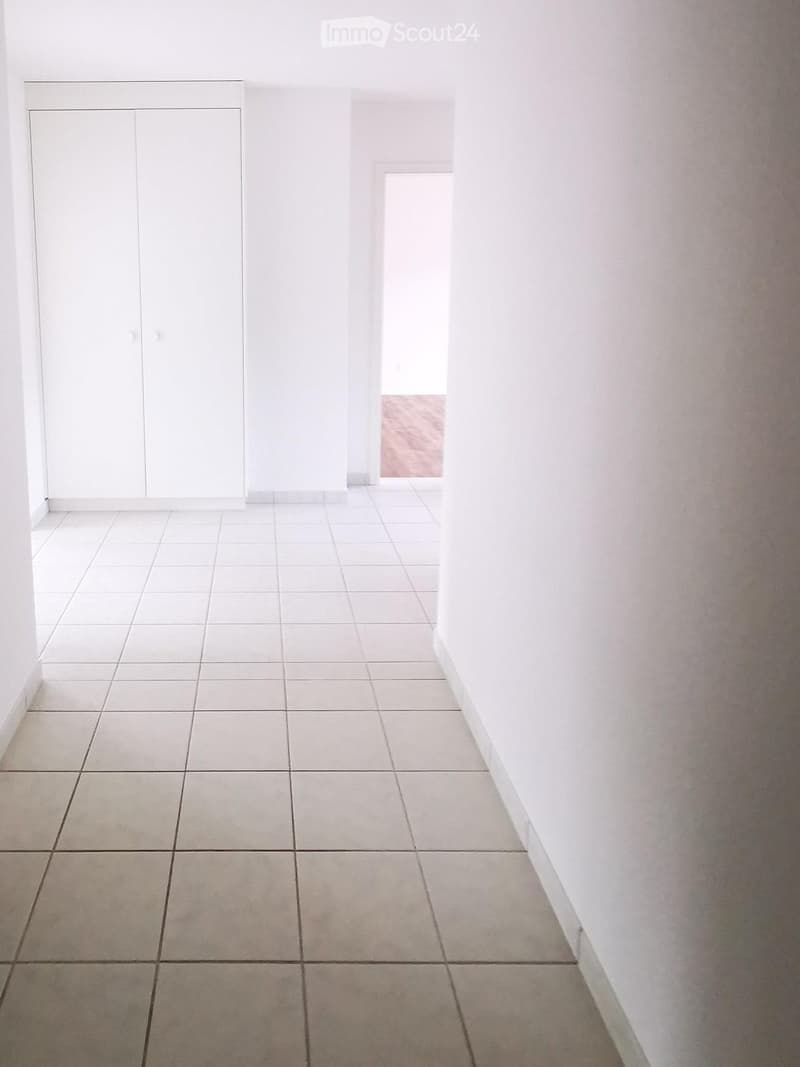 Empty room with white tiled floor and white walls, featuring a white cabinet and a doorway leading to another room.