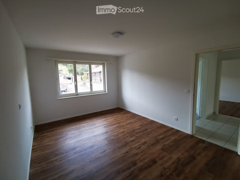 Empty room with wooden flooring, white walls, and a large window with a view of the outside. The room has a door leading to another area with tiled flooring.