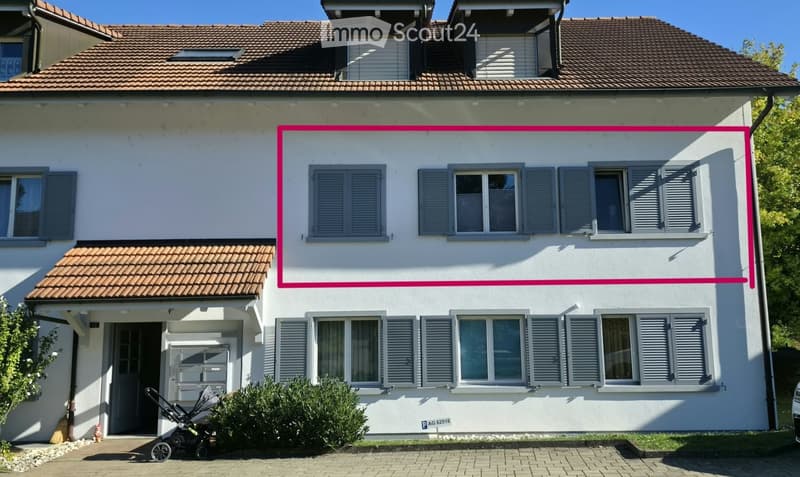 Two-story house with white exterior, grey shutters, and a tiled roof. Features include a small porch with a door, a stroller parked nearby, and a small garden area.