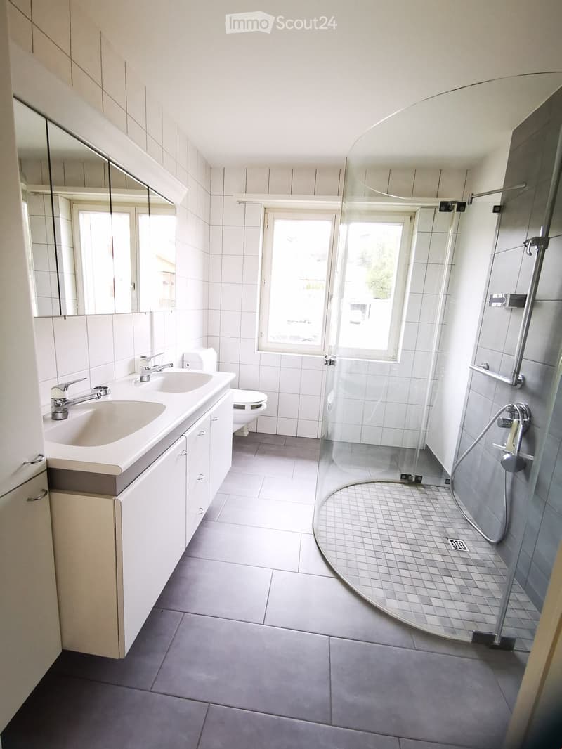Modern bathroom with white tiles, a double sink, a toilet, a shower with glass enclosure, and a large mirror above the sink.