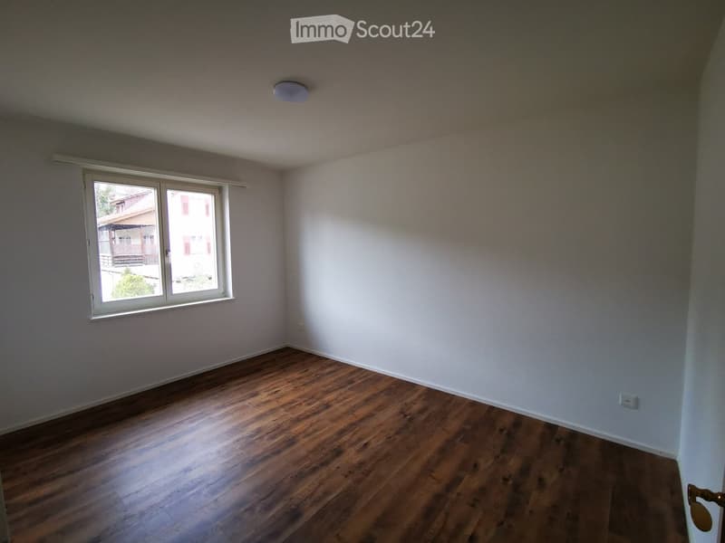 Empty room with wooden flooring, white walls, and a large window with white frame. The room has a single ceiling light fixture and a door visible on the right side.