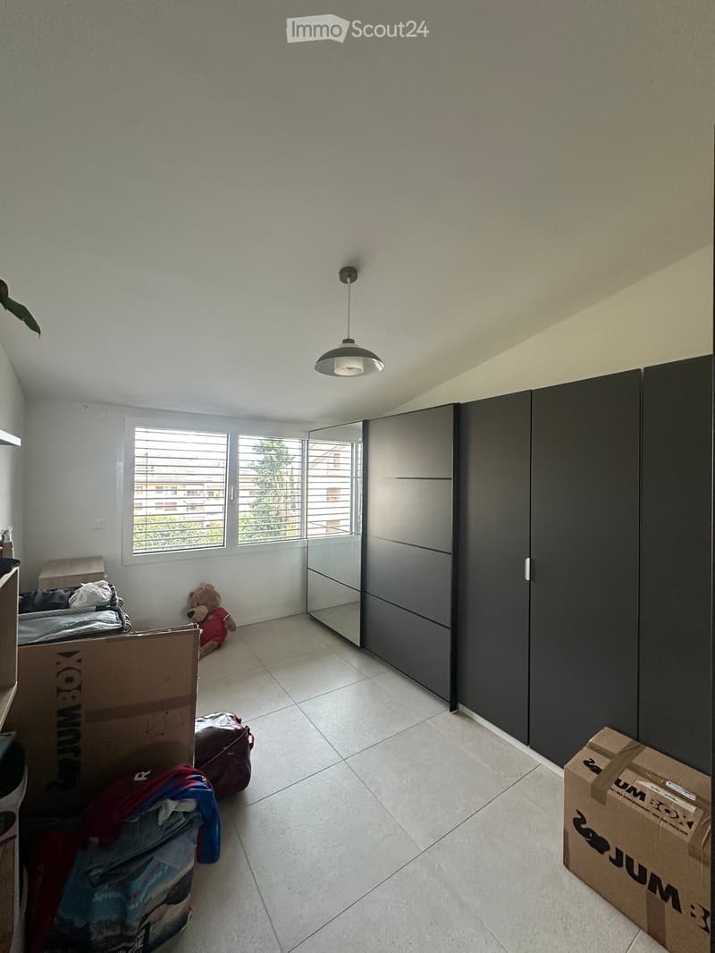 Empty bedroom with a large wardrobe, a bed with bedding, a teddy bear, and several cardboard boxes and suitcases scattered around.