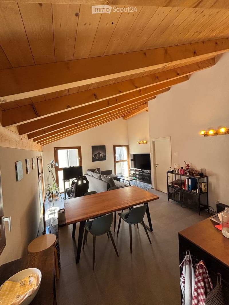A modern, open-concept living and dining area with exposed wooden beams, large windows, and a mix of contemporary furniture including a sofa, dining table, chairs, and a television.