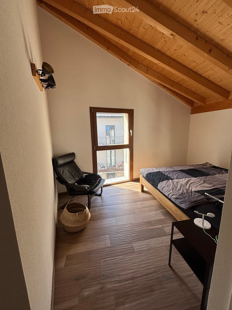 Cozy attic bedroom with wooden slanted ceilings, a bed, a black leather chair, a nightstand with a lamp, and a wicker basket. A large window opens to a balcony.
