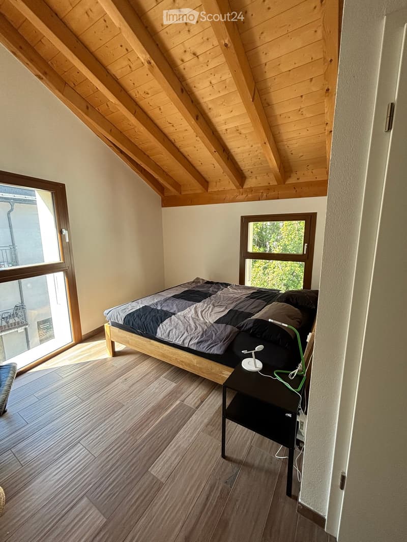 Cozy attic bedroom with wooden slanted ceilings, wooden flooring, and large windows. Features a bed with a dark grey duvet, a nightstand with a lamp, and a balcony access.