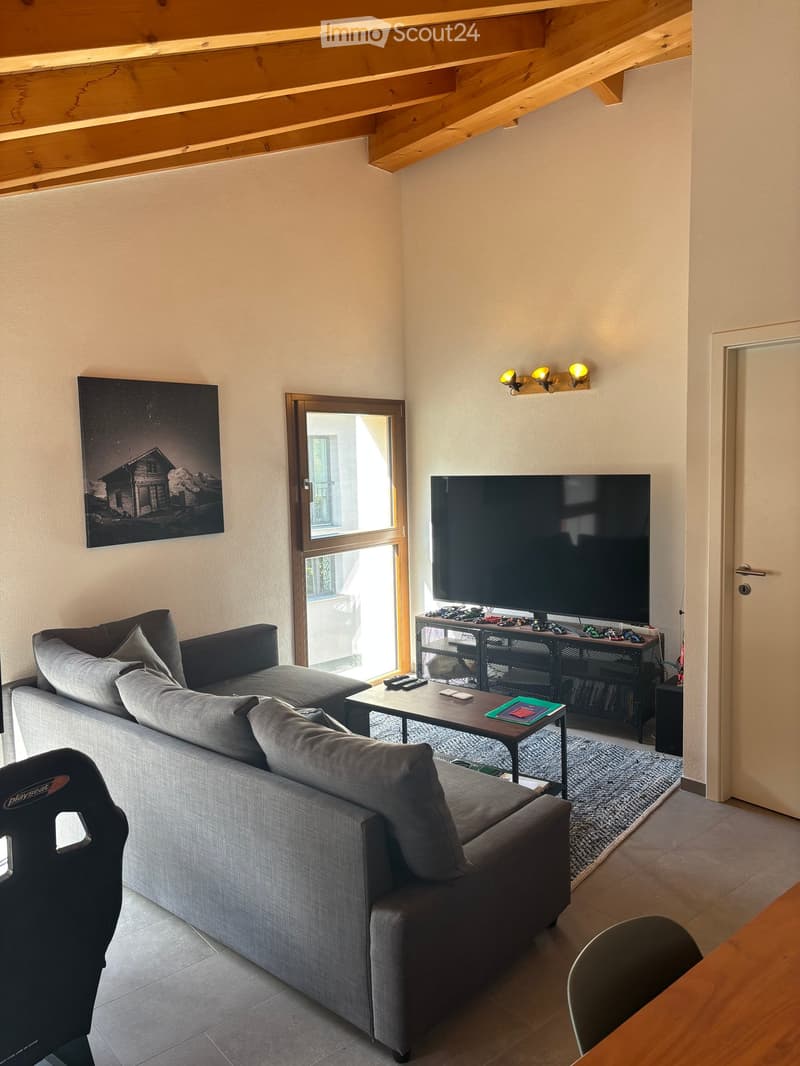 Modern living room with a grey sectional sofa, coffee table, flat-screen TV, and a large window with wooden frame. The room features exposed wooden beams on the ceiling and a cozy rug on the floor.