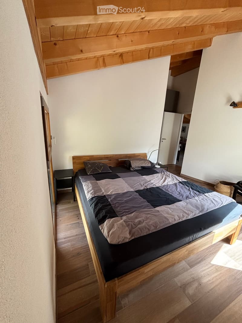 Cozy bedroom with a wooden bed, patterned quilt, nightstand, lamp, and a small chair near the door.