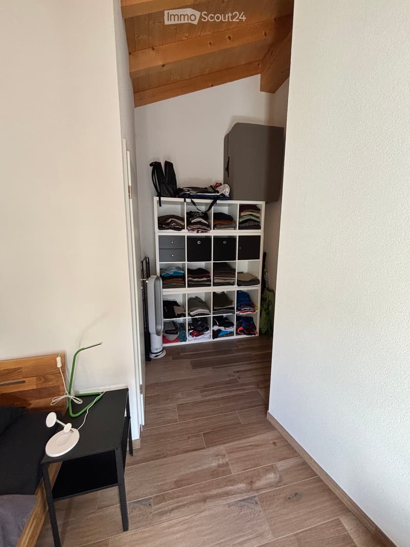 A small hallway with wooden flooring, a black table with a white lamp, a storage unit filled with folded clothes, and a backpack on top.