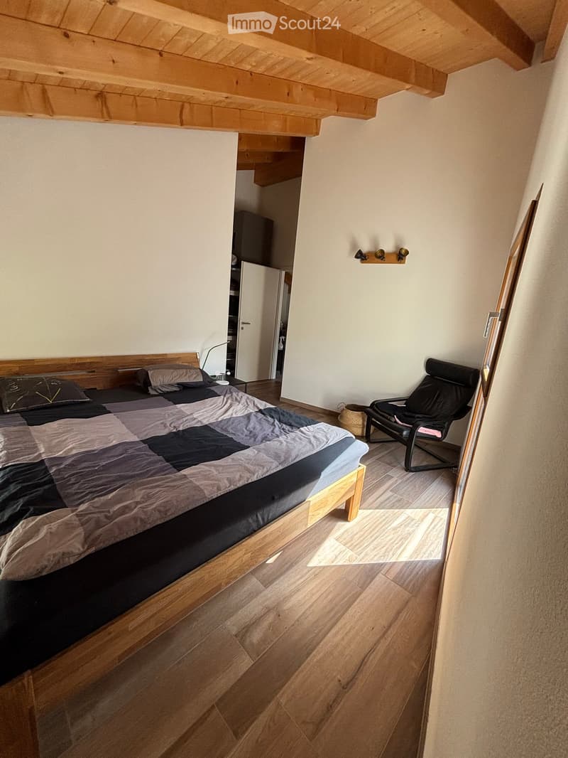 Modern bedroom with wooden flooring, a large bed with a patterned duvet, a black leather chair, and a nightstand with a lamp.
