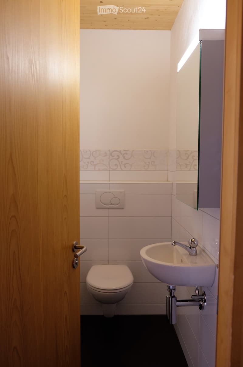 Modern bathroom with white tiles, a toilet, a sink with a chrome faucet, and a mirror above the sink. The walls feature decorative tile borders, and the ceiling is finished with light wood paneling.
