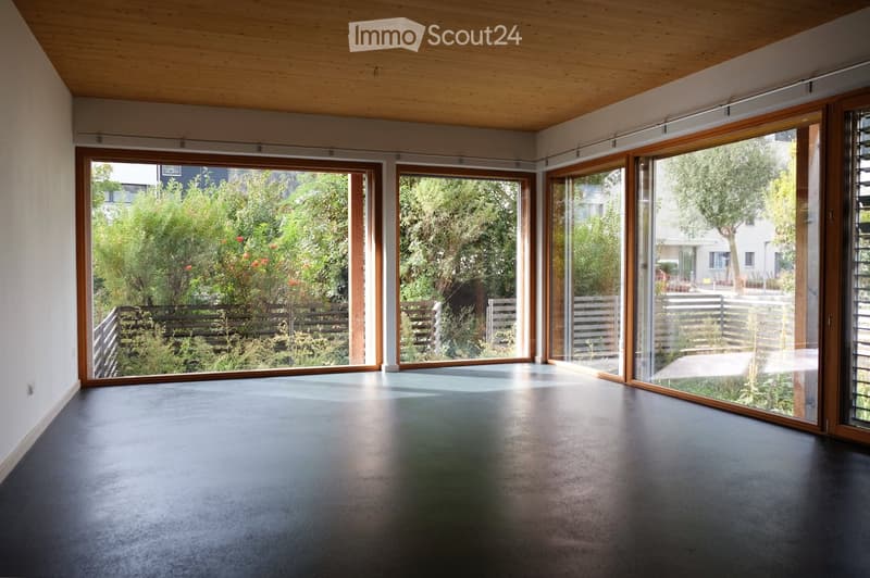 An empty room with a polished concrete floor, large sliding glass doors, and a wooden ceiling. The space is open and bright, with a view of greenery outside.