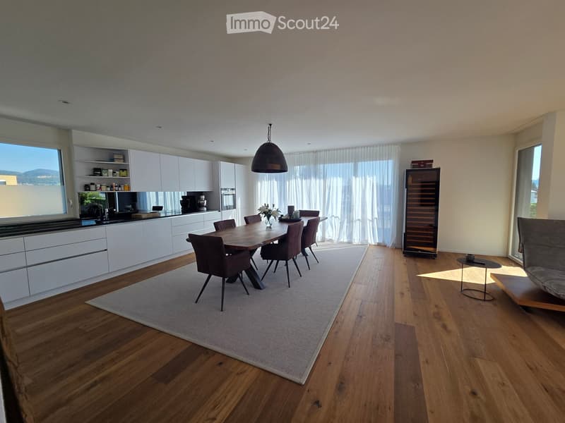Open-plan kitchen and dining area with modern white cabinets, a wooden dining table with chairs, a wine rack, a sofa, and a coffee table. Large windows with curtains allow natural light to fill the space.