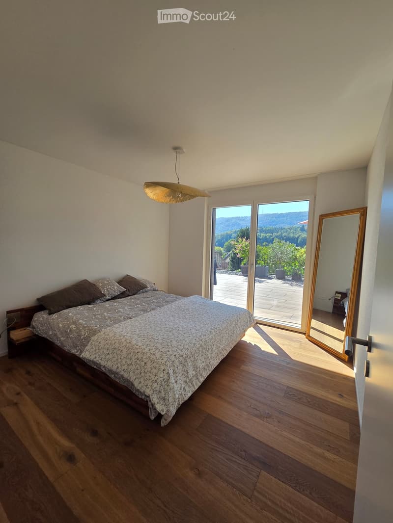 Bright bedroom with wooden flooring, large bed with patterned bedding, nightstand, and a large mirror. Large sliding glass doors open to a scenic outdoor view.