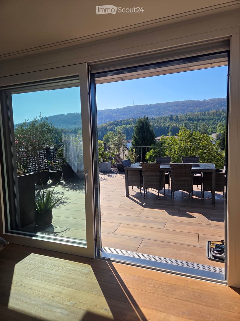 Open sliding glass doors lead to a wooden deck with a dining table and chairs, overlooking a scenic valley and forested hills.