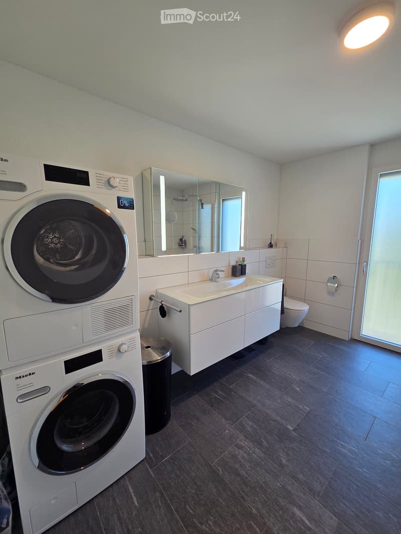 Modern bathroom with a washing machine, sink, mirror, toilet, and a trash can. The room features dark tiled flooring and white cabinetry.