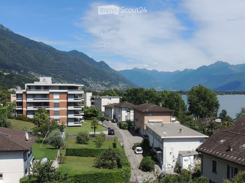 A multi-story residential building with balconies and surrounding greenery, set against a backdrop of mountains and a lake.