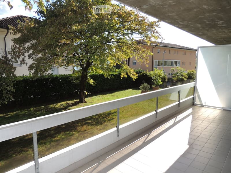 Balcony with tiled floor, white railing, and glass panels overlooking a well-maintained garden with a tree, shrubs, and neighboring buildings.