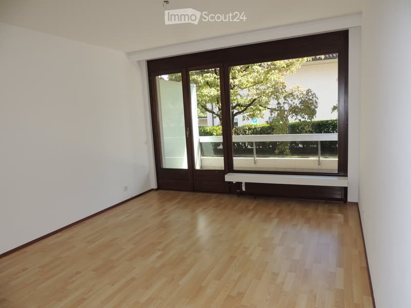 Empty room with light wooden flooring, white walls, large sliding glass doors leading to a balcony, and a white bench beneath the windows.