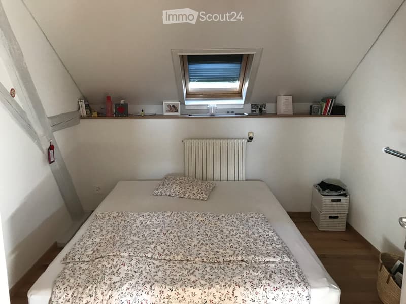 Cozy attic bedroom with a single bed, patterned bedding, a small window, a radiator, and a shelf with books and decorative items.