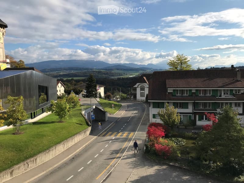 A quiet suburban street lined with houses, trees, and a modern building on the left, with a scenic mountain backdrop.