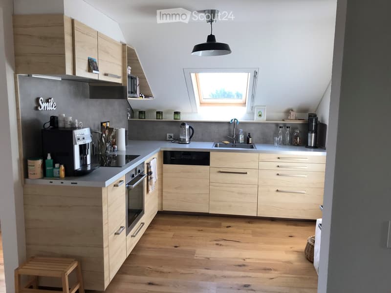 Modern kitchen with light wooden cabinets, stainless steel appliances, and a central island. Features include a coffee maker, oven, dishwasher, and a hanging pendant light.