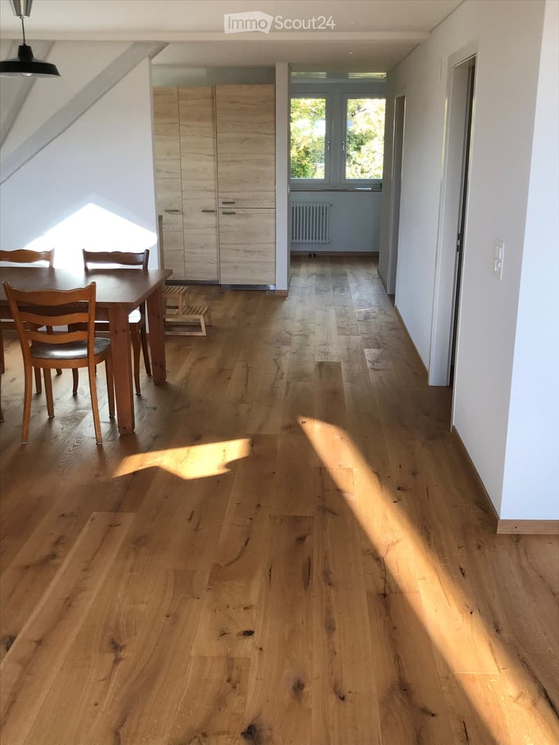 Bright, airy room with wooden flooring, a dining table with chairs, and a large wardrobe. Sunlight streams in from a window at the end of the room.