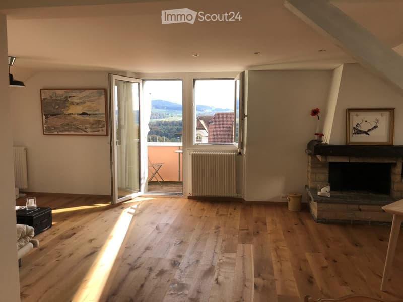 Bright living room with wooden flooring, fireplace, large windows offering a scenic view, and modern decor including a sofa, small table, and artwork.