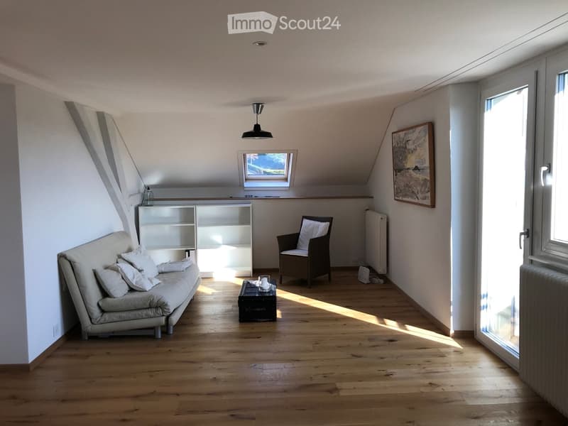 Bright, airy attic room with wooden flooring, white walls, and large windows. Features a sofa, armchair, small table, shelf, and artwork.