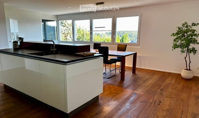 Modern kitchen with white cabinets, black countertop, island, sink, and large windows overlooking a green view. Includes a dining area with a table and chairs, a blender on the counter, and a potted plant in the corner.