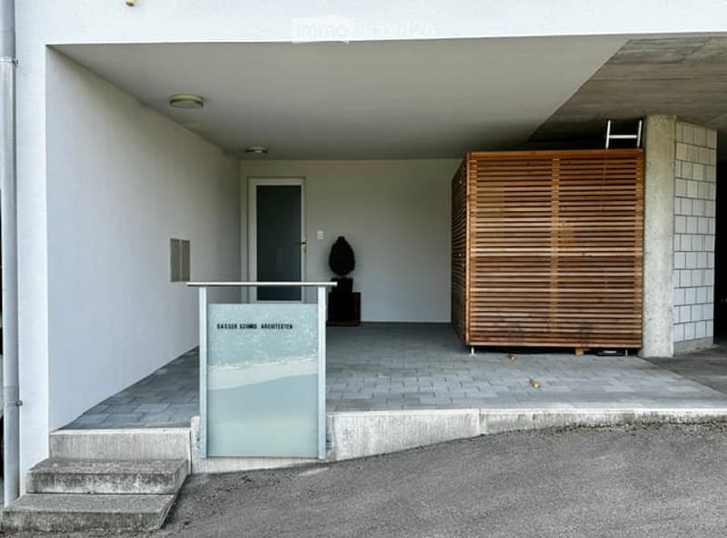 Modern building entrance with raised ground floor, concrete steps, glass panel, wooden screen, and small sculpture.