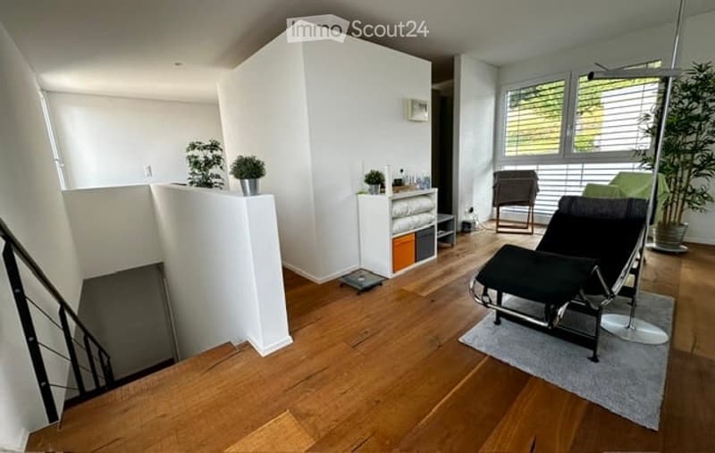 Modern, minimalist living room with wooden flooring, white walls, and large windows. Features a black leather sofa, a matching chair, a small table with plants, and a storage unit with shelves.