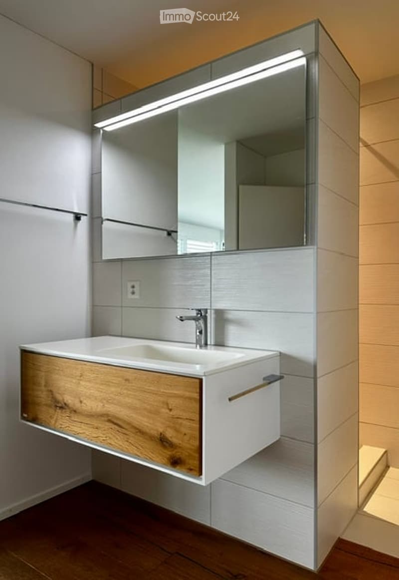 Modern bathroom with a sleek, floating vanity in light wood and white ceramic, a large mirror with integrated LED lighting, and a chrome faucet.