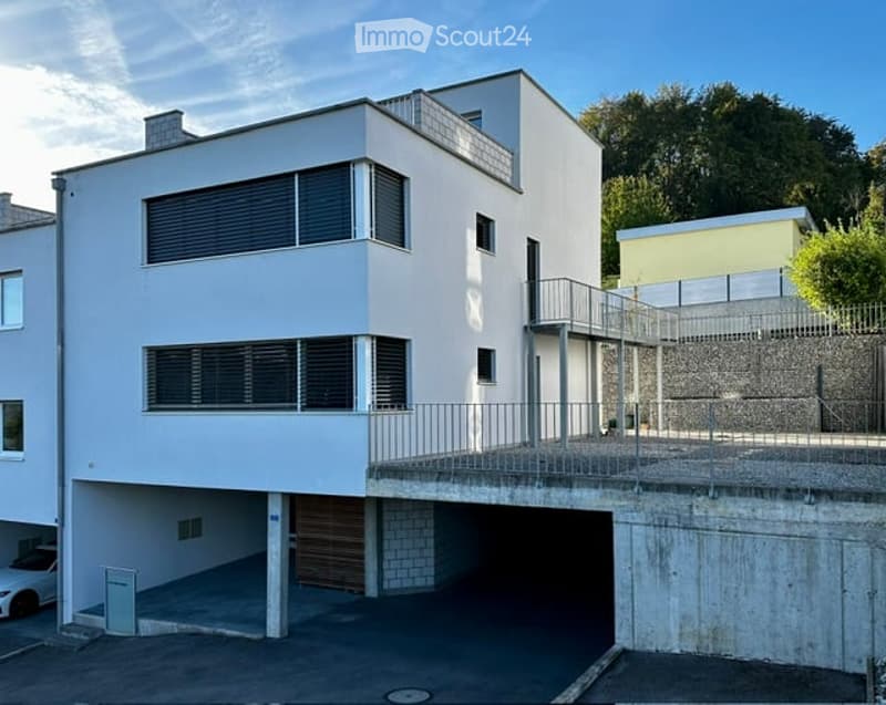 Modern two-story building with white facade, large windows, and a covered garage area below. Features a raised ground floor and a balcony on the upper level.