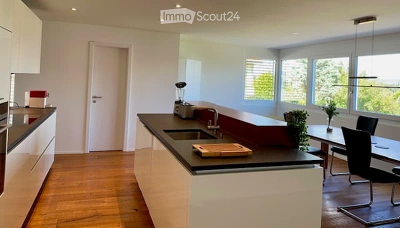 Modern kitchen with white cabinets, black countertops, and a central island. Features include a blender, cutting board, and a small dining area with chairs and a table.