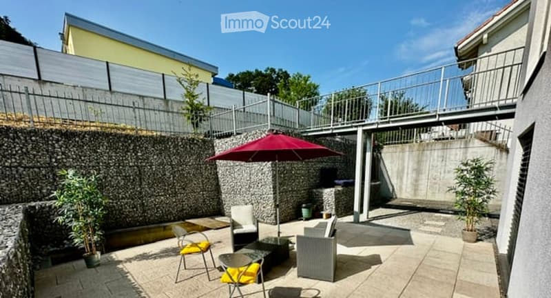 Patio area with outdoor furniture including chairs, a table, and an umbrella, surrounded by a stone wall and greenery.