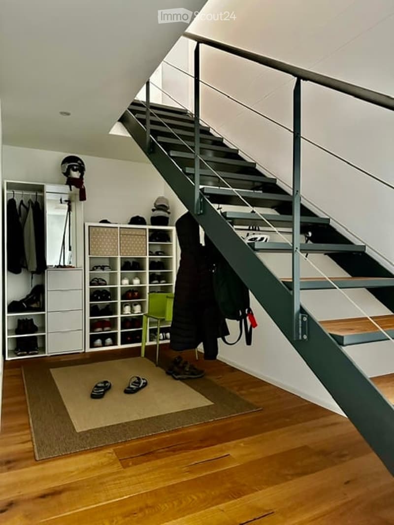 A modern hallway with a wooden floor, a staircase with metal railings, a coat rack with jackets, a shoe rack, a small green stool, and storage boxes.
